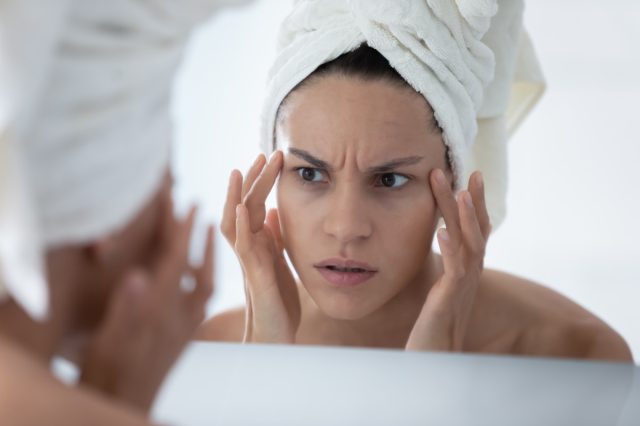 Unhappy woman wearing white bath towel checking skin after shower, looking in mirror, touching facial skin.
