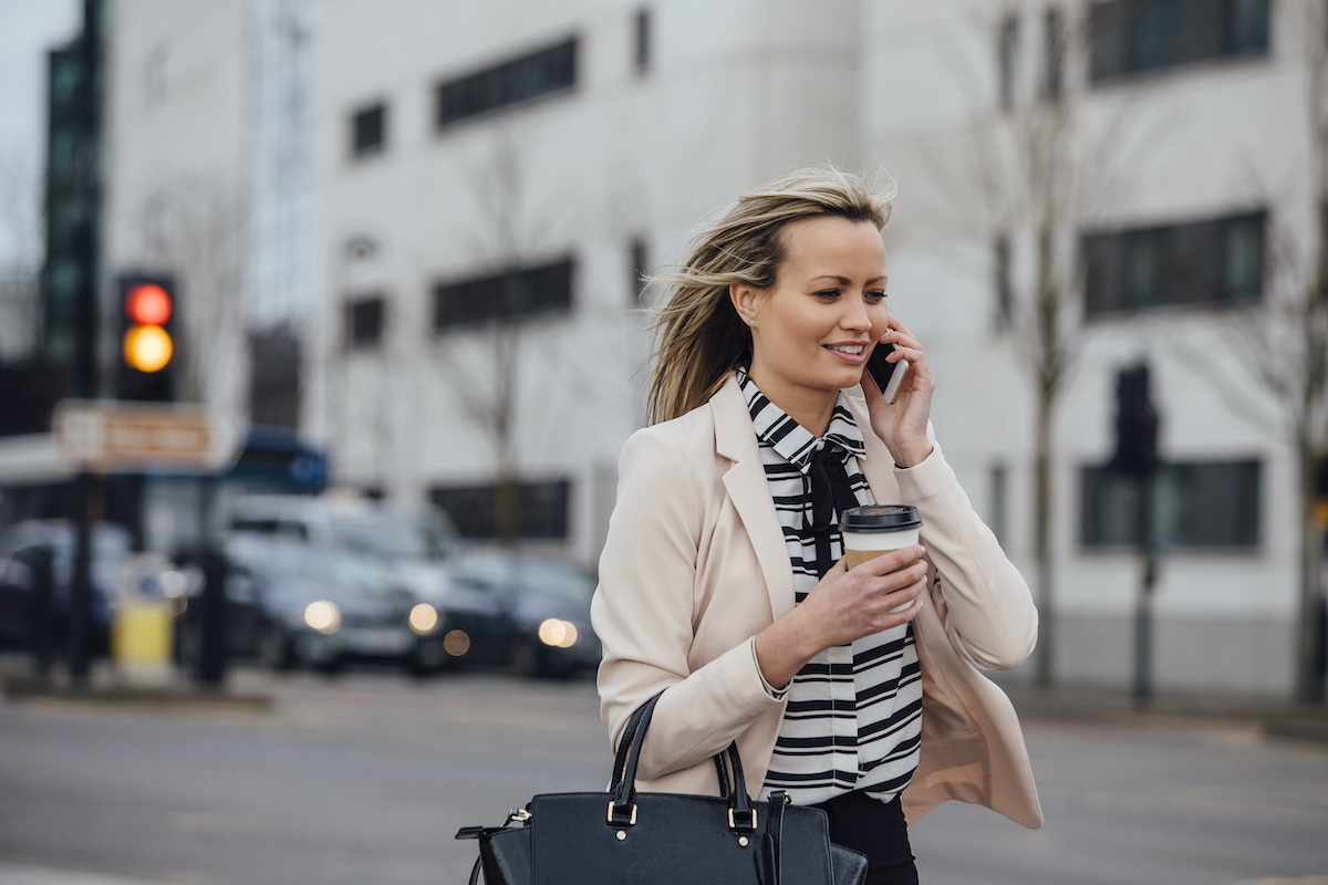 Businesswoman is walking to work in the city. She is holding a coffee cup and is talking on the phone.