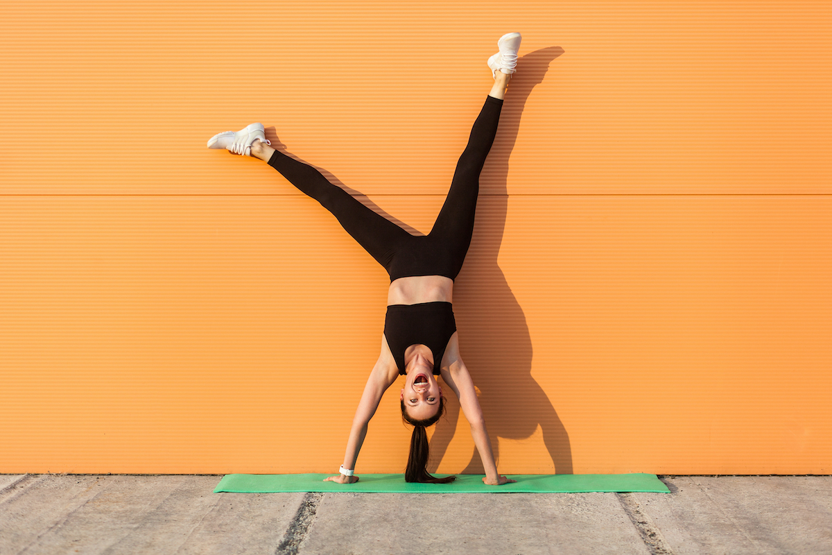 Overjoyed excited girl with perfect athletic body in tight sportswear doing yoga handstand pose against wall and laughing, shouting from happiness. Gymnastics for body balance outdoor workouts