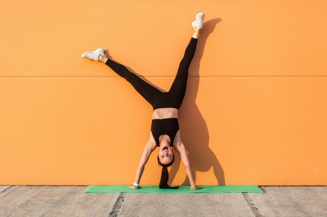 Overjoyed excited girl with perfect athletic body in tight sportswear doing yoga handstand against wall and laughing, screaming with happiness.  Gymnastics for body balance outdoors