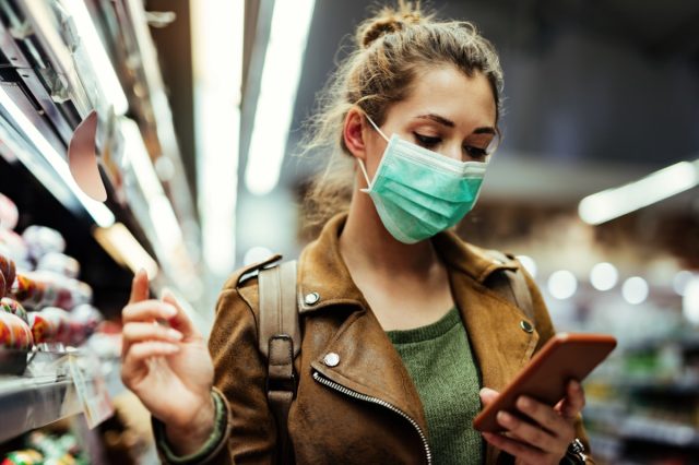 Young woman wearing protective mask on her face and reading shopping list on mobile phone in grocery store during virus pandemic