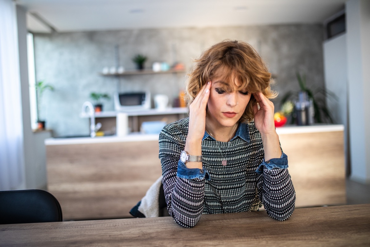 Woman suffering from headache, holding head with hand while sitting in dinning room at table.