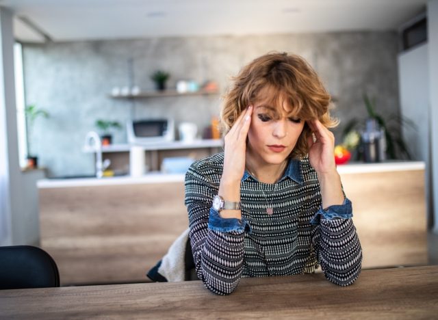 Woman suffering from headache, holding head with hand while sitting in dinning room at table.