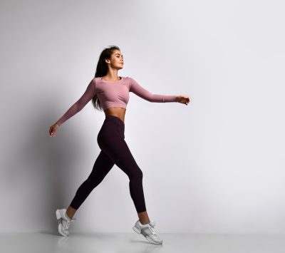 Young athletic woman brunette girl in a good shape with hair in a ponytail in trendy sportswear gym uniform jumps dances does exercises workout isolated on gray background walking