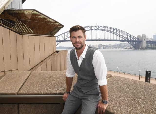 chris hemsworth in gray vest, gray pants, and white shirt sitting on a ledge at sydney opera house
