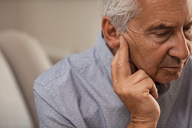 Страничен изглед на старши мъж със симптом на загуба на слуха.  Зрял мъж, седнал на дивана с пръсти близо до ухото, страдащ от болка.