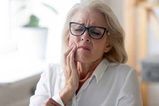 Unhappy mature woman in glasses touching her jaw.