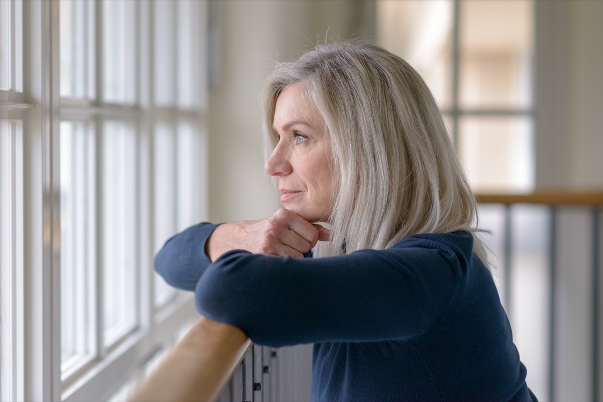 Sad blond woman watching through a window with a serious expression resting her chin on her hands as she leans on a wooden railing