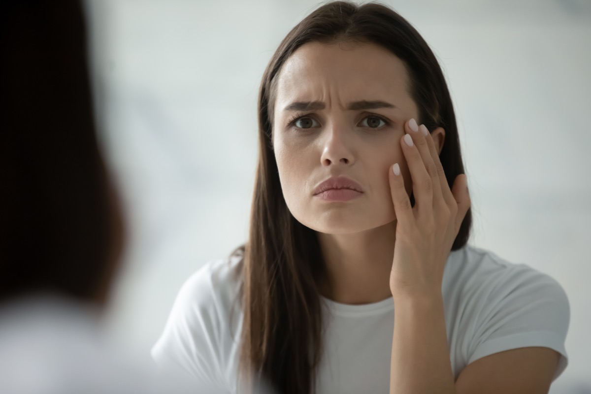 Unhappy young woman wearing white t-shirt checking skin, looking in mirror, standing in bathroom.