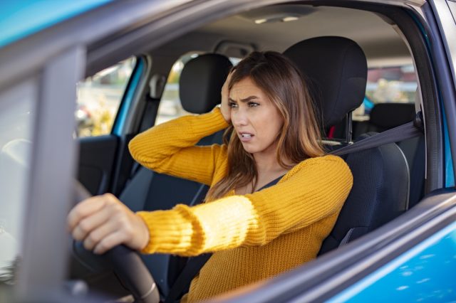 Female driver sits at wheel in car, touches her head.