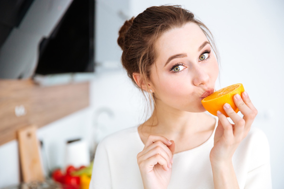woman in white top standing in kitchen eating orange