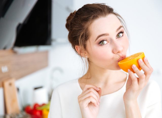 woman in white top standing in kitchen eating orange