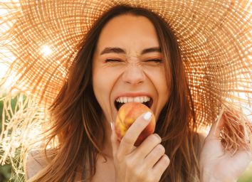 woman standing outdoors while eating a peach and wearing a straw hat
