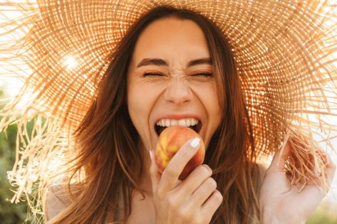 woman standing outdoors while eating a peach and wearing a straw hat