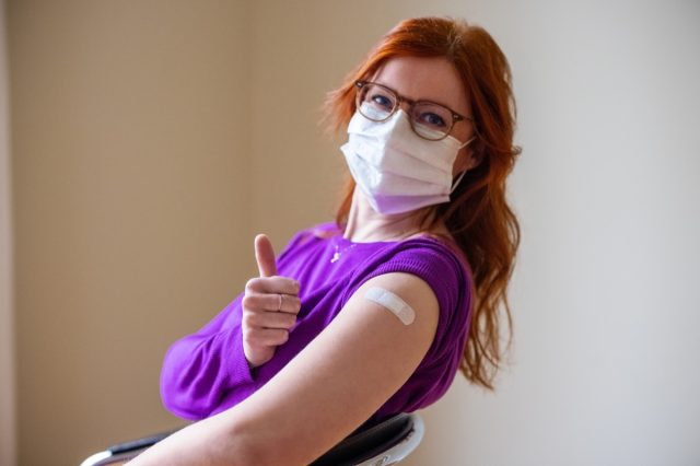 A woman in a mask looks at the camera and shows a thumbs up after receiving a vaccine against COVID-19.