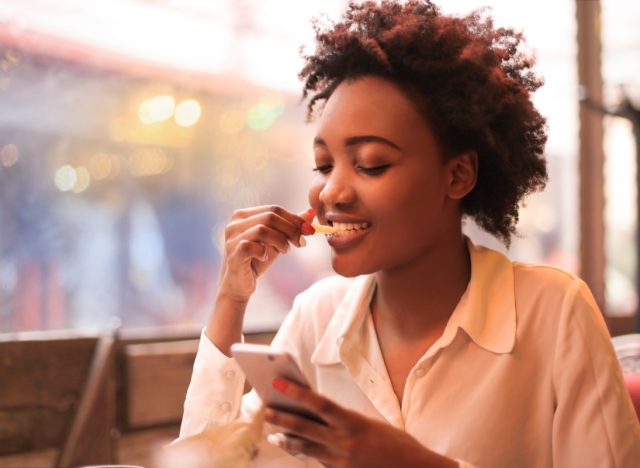 young woman looking at phone and eating fries
