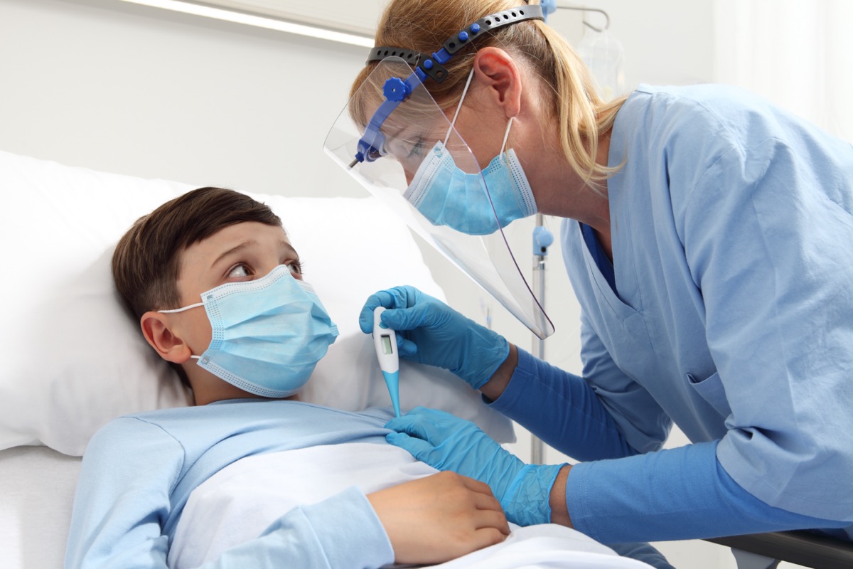 A nurse with a thermometer measures fever on a child patient in a hospital bed, wearing a protective visor and surgical mask.