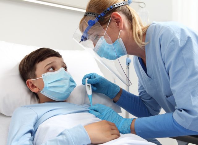 Nurse with thermometer measures fever on patient child in hospital bed, wearing protective visor and surgical mask.
