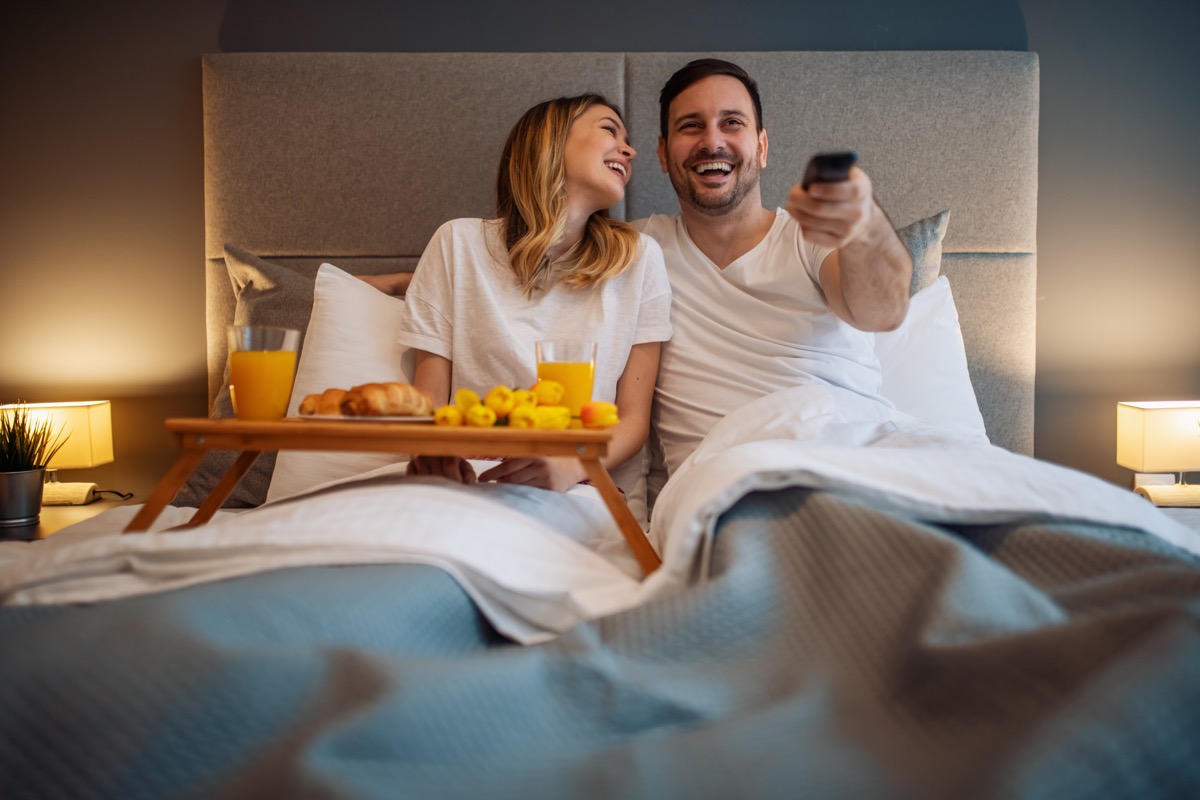 30-something couple eating breakfast in bed and watching tv
