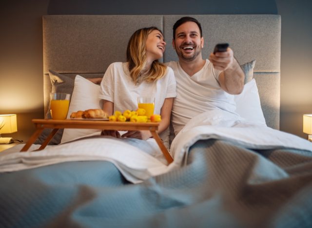 30-something couple eating breakfast in bed and watching tv