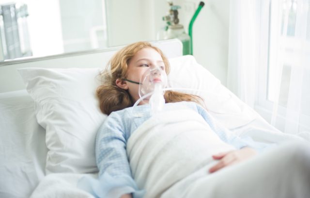 Little girl is sick using oxygen mask on her face laying in bed at hospital.