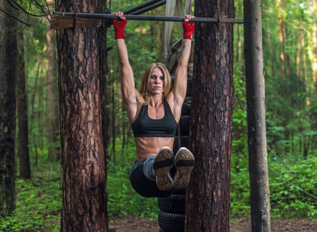 Fit woman doing hanging leg lifts abs muscles exercise on horisontal bar working out outside.