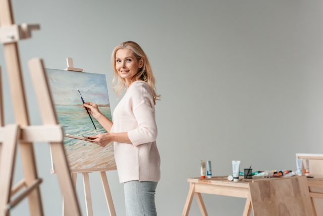 mature female artist smiling at camera while painting on easel in art studio