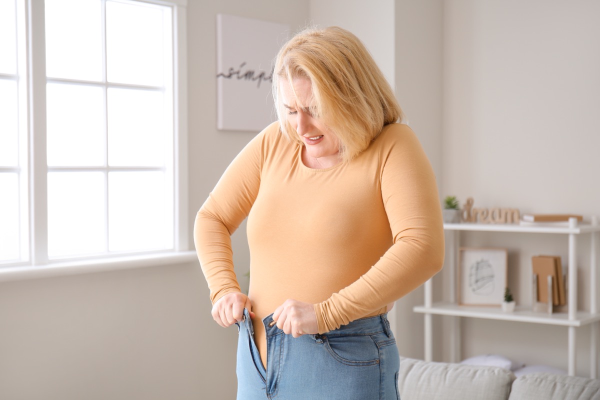 Overweight woman in tight clothes at home is trying to fit into tight jeans.
