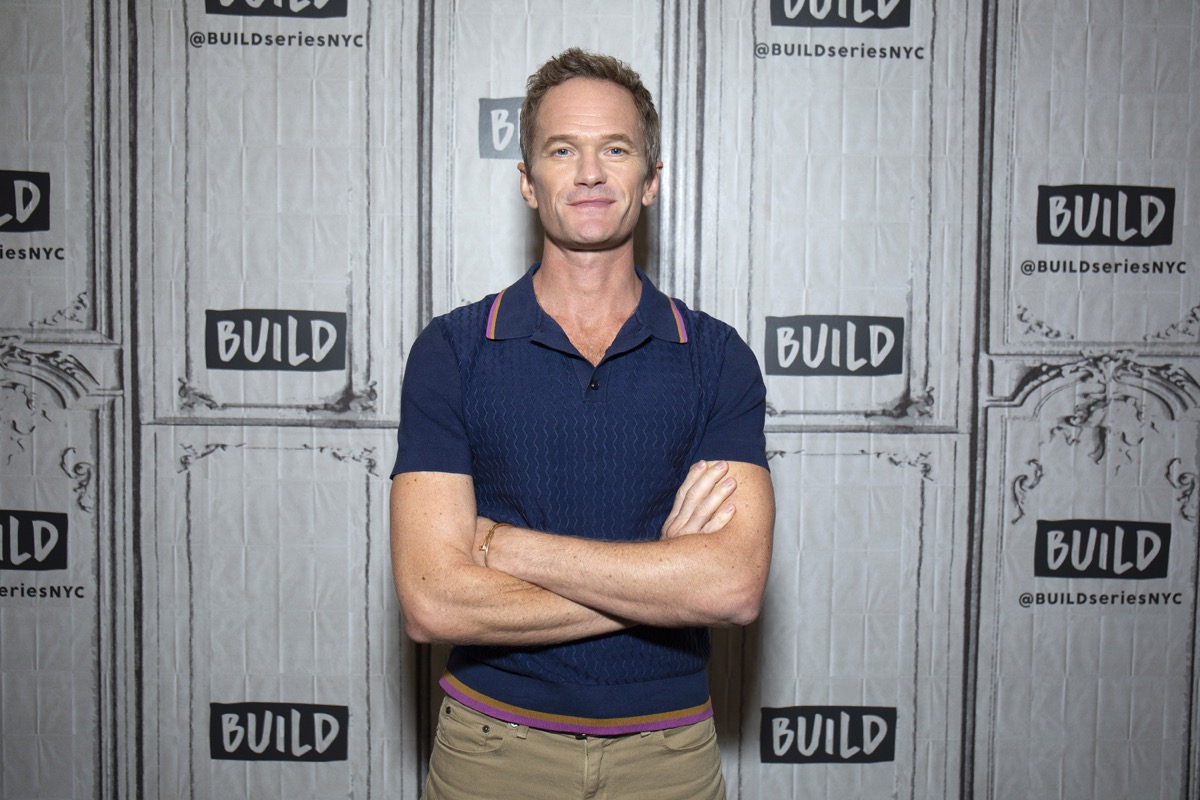 neil patrick harris with arms crossed wearing blue polo shirt in front of build studios step and repeat