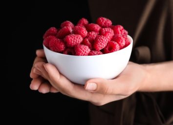 hands holding white bowl of raspberries against dark background