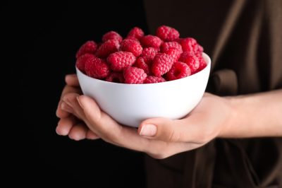hands holding white bowl of raspberries against dark background