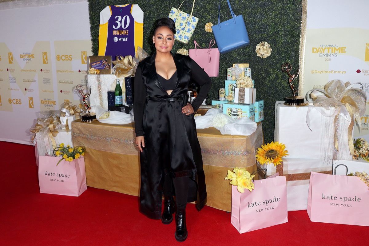 raven-symone in long black satin dress or cape in front of gifting suite and pink kate spade bags