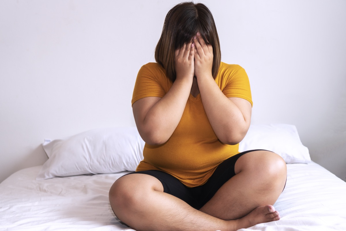 Overweight young woman sitting on white bed while holding hands cover on her face at home. Upset female suffering from extra weight in the bedroom. Obesity unhealthily concept.