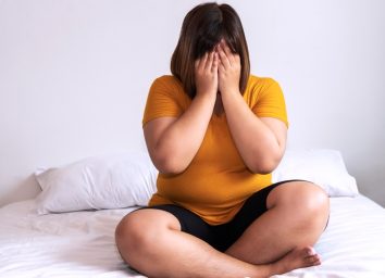 Overweight young woman sitting on white bed while holding hands cover on her face at home. Upset female suffering from extra weight in the bedroom. Obesity unhealthily concept.
