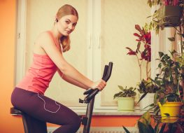 Active young woman working out on exercise bike stationary bicycle. Sporty girl training at home listening music. Fitness and weight loss concept.