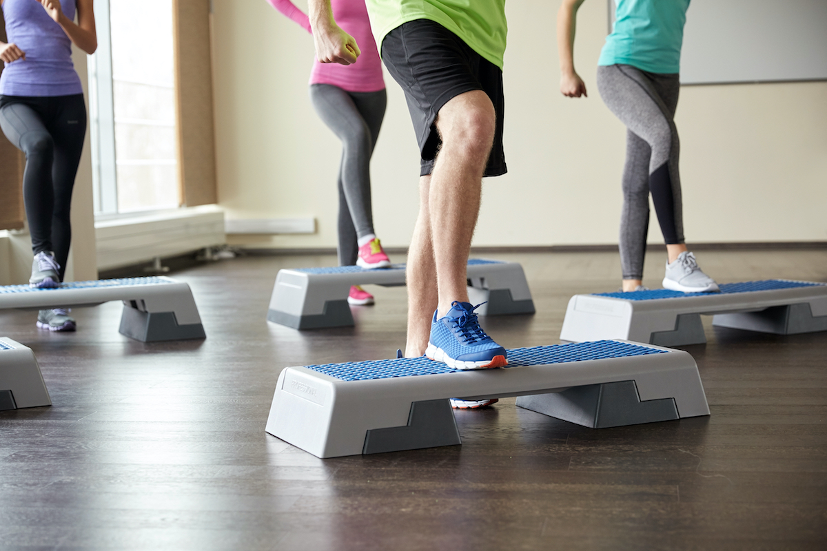 fitness, sport, aerobics and people concept - group of smiling people working out and flexing legs on step platforms in gym