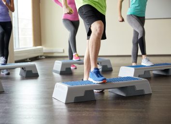 fitness, sport, aerobics and people concept - group of smiling people working out and flexing legs on step platforms in gym