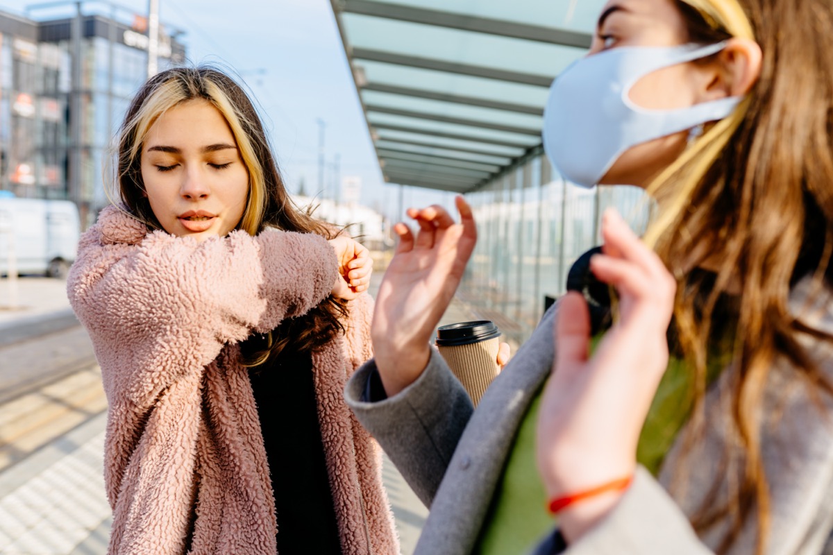 Young sick student teenager woman outside at bus stop is sneezing into the elbow by an allergy or cold. Scared woman in protective mask afraid cough woman outdoor