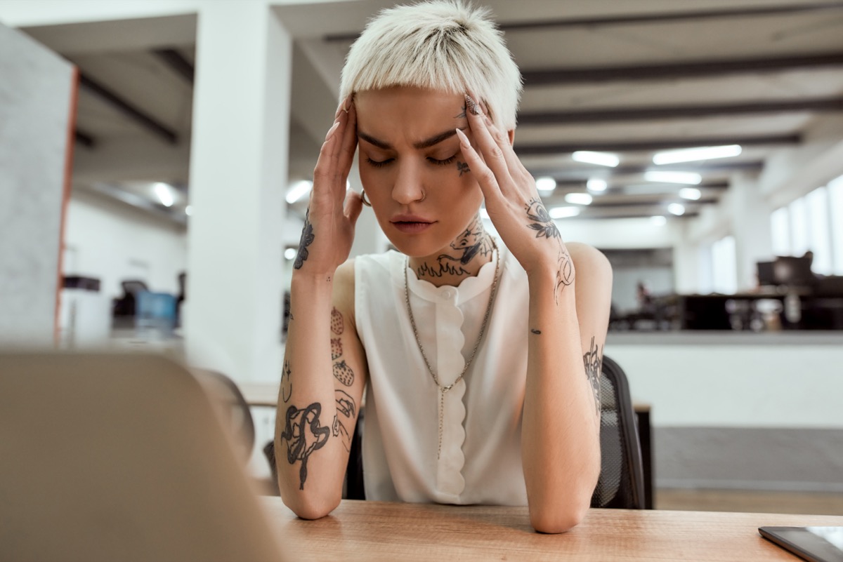 Exhausted young tattooed business woman keeping eyes closed, touching head and suffering from the headache while sitting at her working place in the modern office