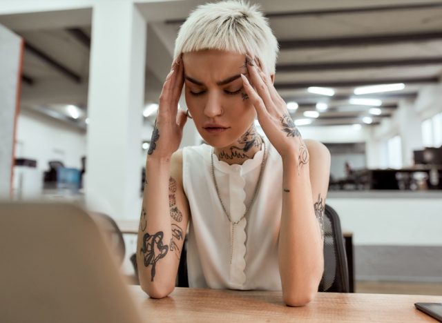 Exhausted young tattooed business woman keeping eyes closed, touching head and suffering from the headache while sitting at her working place in the modern office