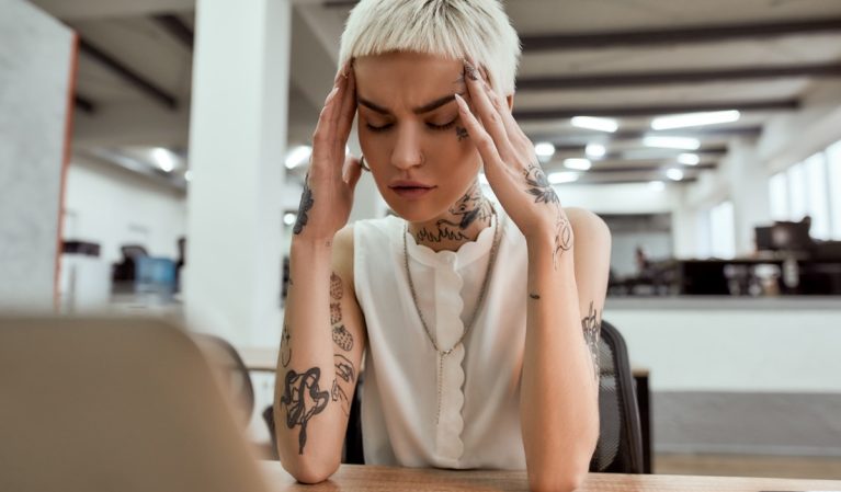 Exhausted young tattooed business woman keeping eyes closed, touching head and suffering from the headache while sitting at her working place in the modern office