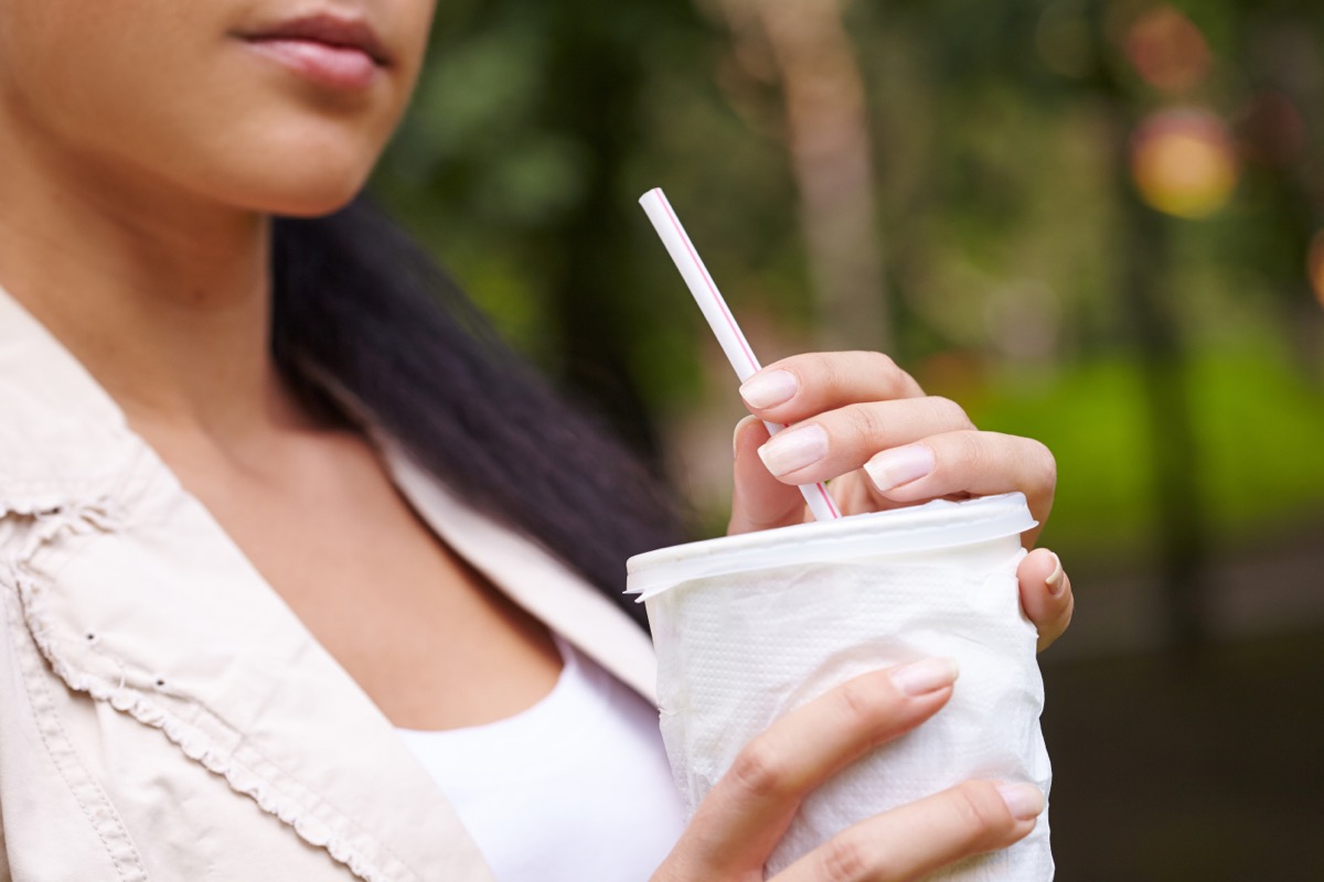 young woman drinking from white cup with straw while sitting oudoors