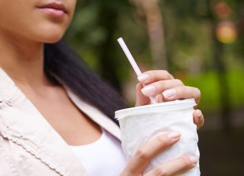 young woman drinking from white cup with straw while sitting oudoors