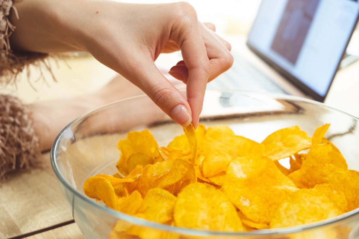 woman in furry sweater reaching into clear glass bowl to grab potato chip while laptop sits in background