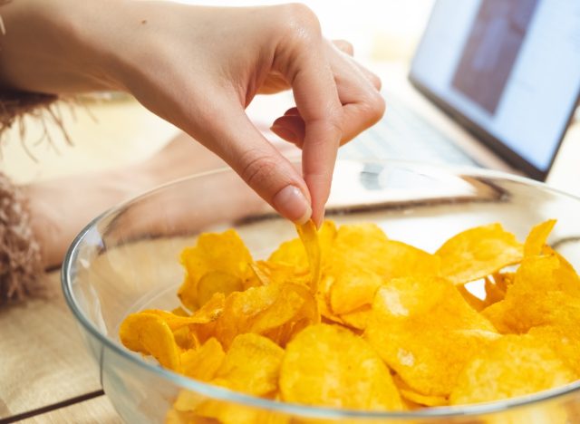 woman in furry sweater reaching into clear glass bowl to grab potato chip while laptop sits in background