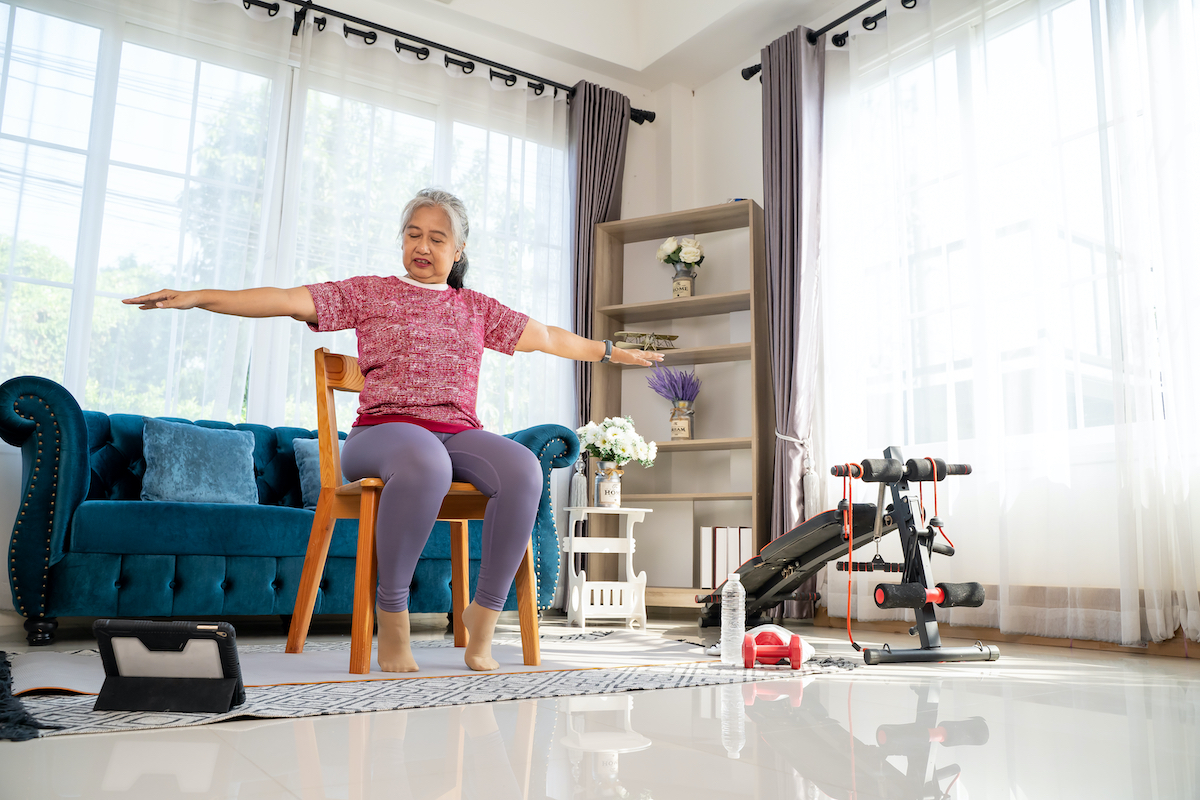 Senior woman sitting on chair and spread both arms and jog in the side to stretch the arm muscles, for good healthy with training exercise online on tablet, workout online from home concept