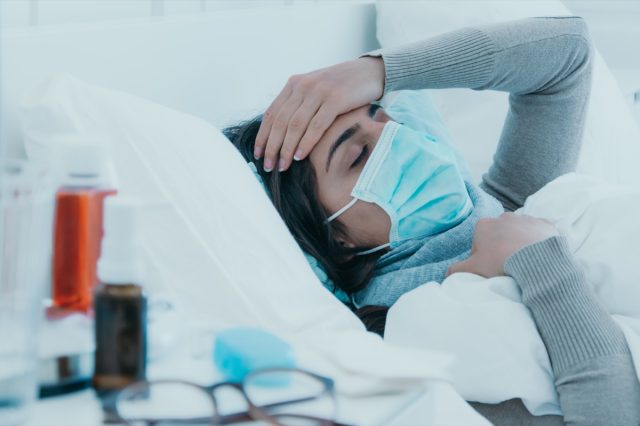 Млада болна жена лежи уморена в леглото с маска за лице и държи главата си от главоболие.