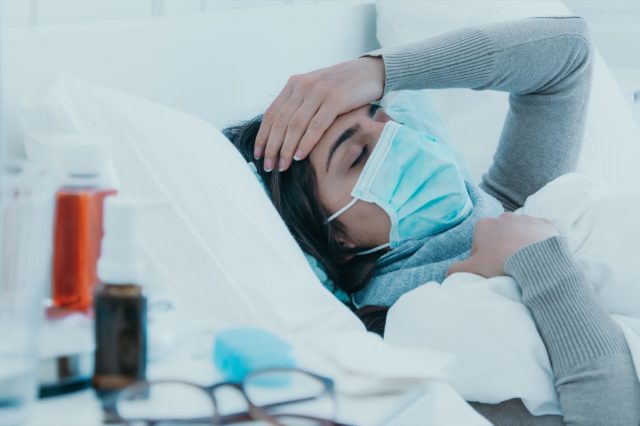 Young sick woman lies tired in bed with a face mask and holds her head because of a headache.