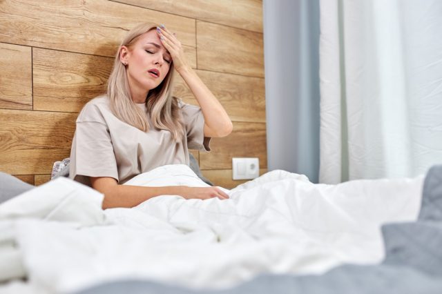 Frau, die an Grippe erkrankt ist und allein zu Hause auf ihrem Bett sitzt, eine hohe Temperatur oder eine hohe Temperatur hat und ihre Stirn berührt