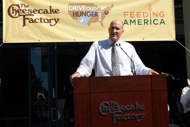 bald man standing at podium outdoors in front of cheesecake factory and feeding america banner